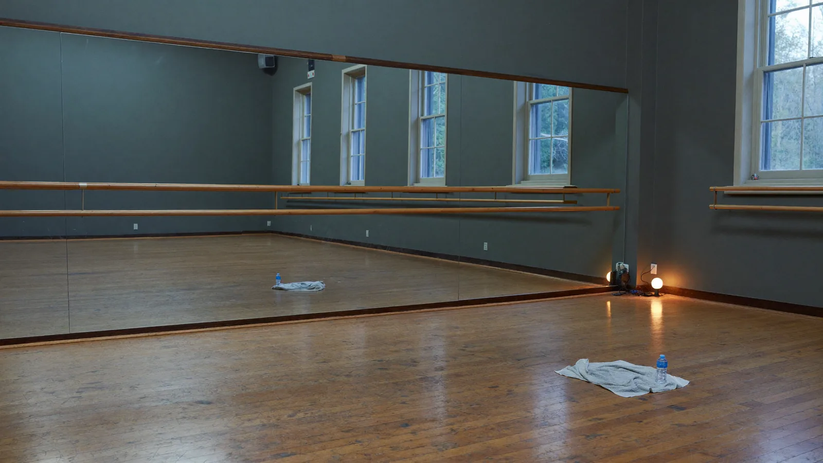 An empty dance studio at dusk: mirror wall, wooden barre, a towel and water bottle left on the floor, one lamp on